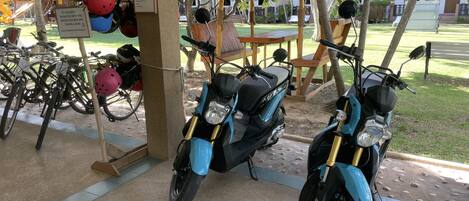 A covered parking area with two blue scooters, helmets, and a sign indicating registration for bike rental.