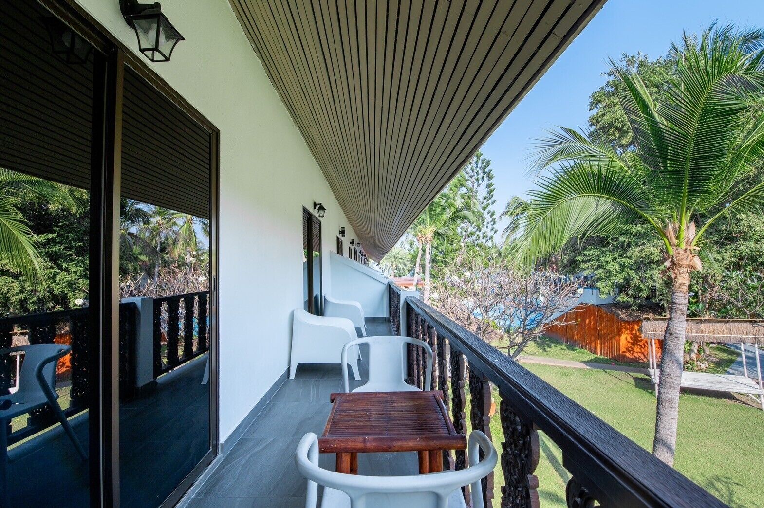 A balcony with a wooden table and white chairs, a wooden railing, and a view of a pool and palm trees.