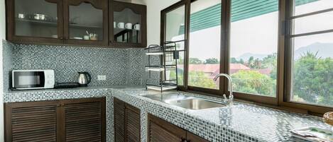 Una cocina con armarios de madera oscura, salpicadero de azulejos, microondas, fregadero y ventana con vistas a zonas verdes y edificios.