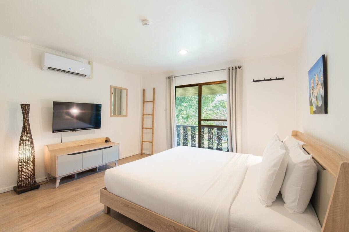 A bedroom with a bed, a TV, a dresser, a ladder, and a balcony with a view of greenery.
