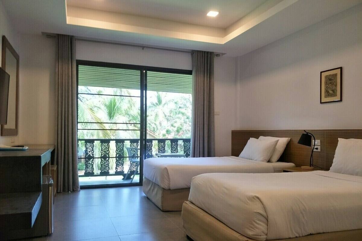 A hotel room with two beds, a desk, a chair, and a balcony with a view of greenery.