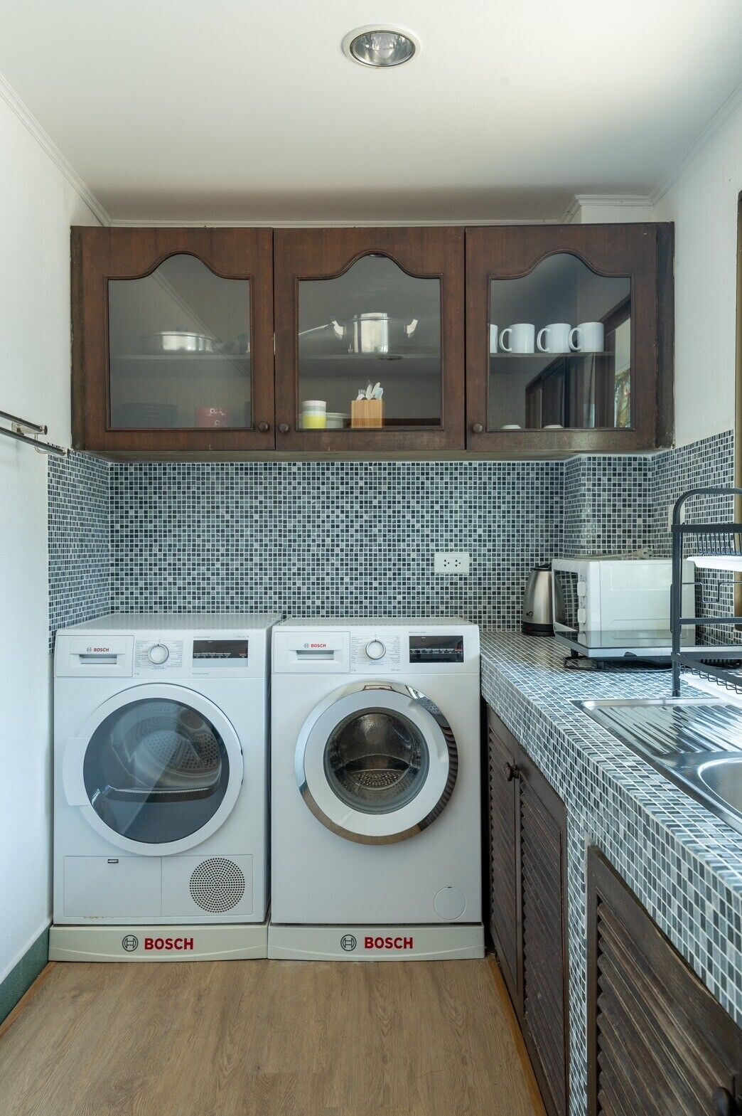 A compact kitchen with two Bosch washing machines, a tiled backsplash, wooden cabinets, and a stainless steel sink.