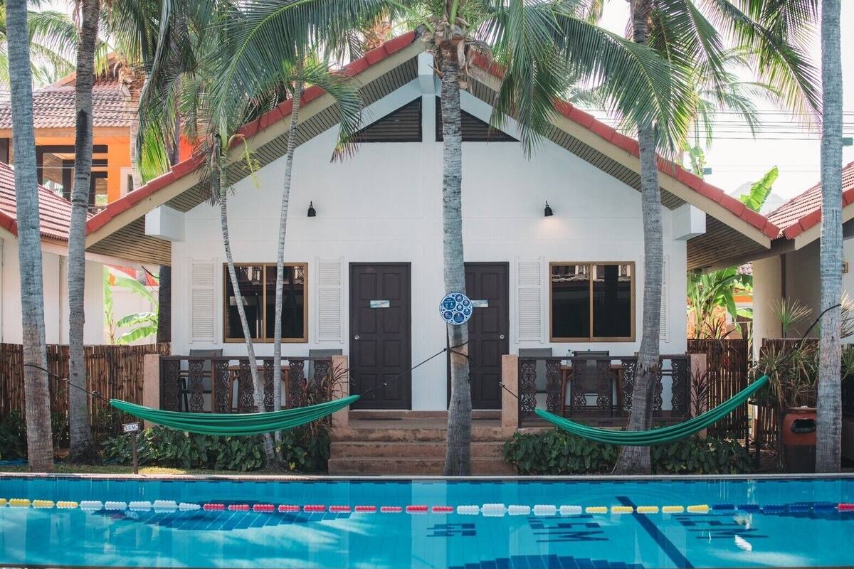 A white building with a red roof, two doors, and windows, surrounded by palm trees and a swimming pool.