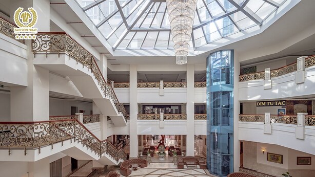 Ein mehrgeschossiges Atrium mit Sitzgelegenheiten in der Mitte, einer Kandelaber-Leuchte und einem gläsernen Aufzugsschacht.