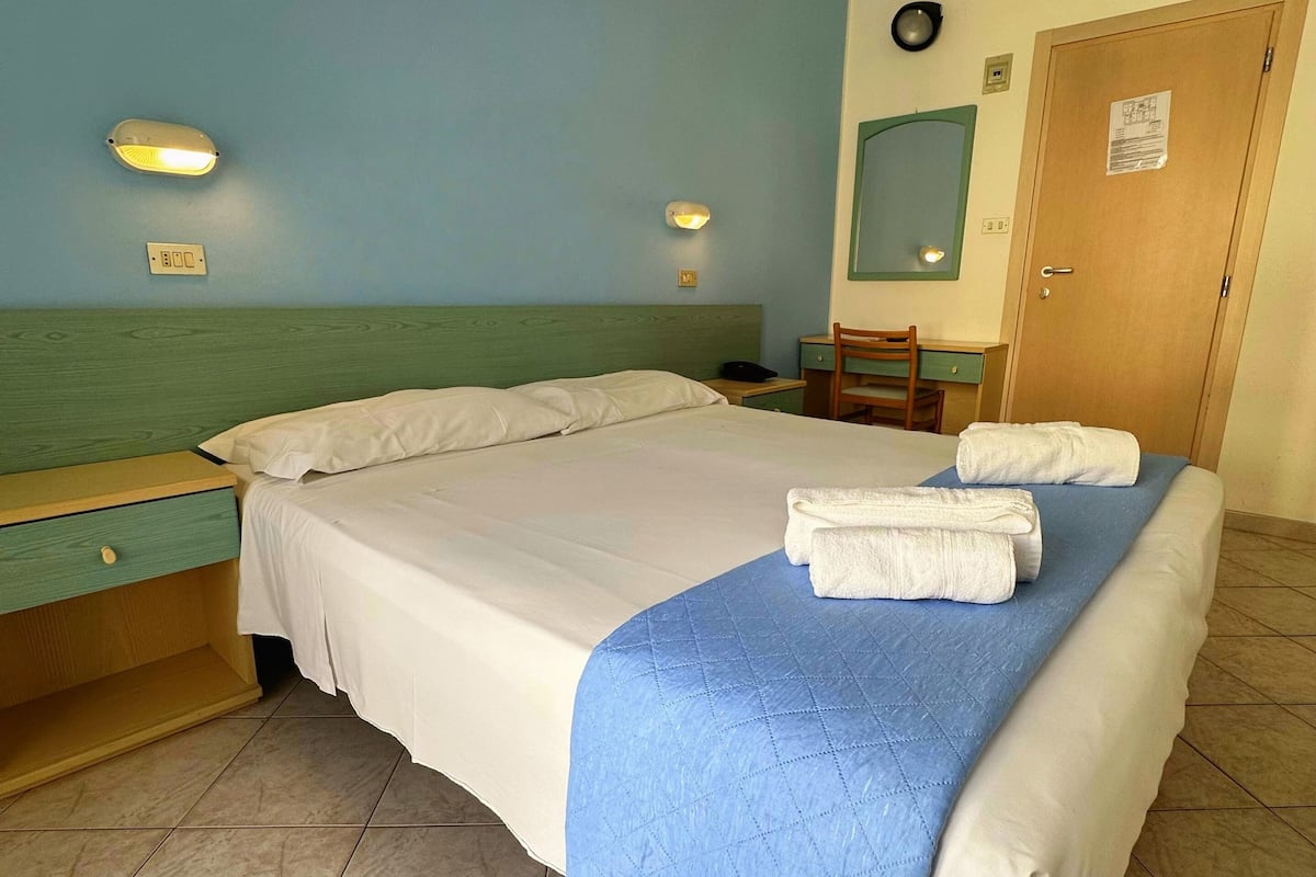 A hotel room with a bed, bedside table, wall-mounted lamp, and a door with a notice.