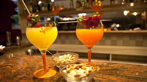 Two cocktails with strawberries on the rim, a bowl of nuts, and a bar counter with bottles in the background. - Hotel Sant'Angelo (Riccione)