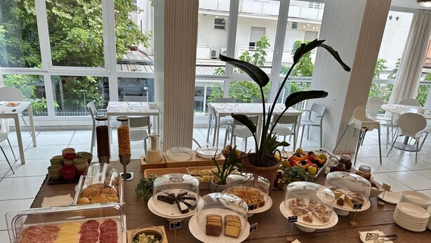 A buffet setup with various food items, a large window with a view of buildings, and indoor plants. - Hotel Sant'Angelo (Riccione)
