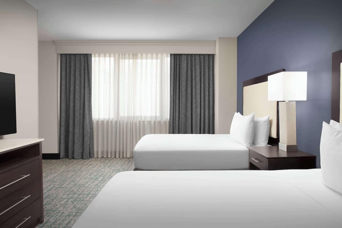 Una habitación de hotel con cama, mesita de noche, lámpara, televisión y ventana con cortinas.