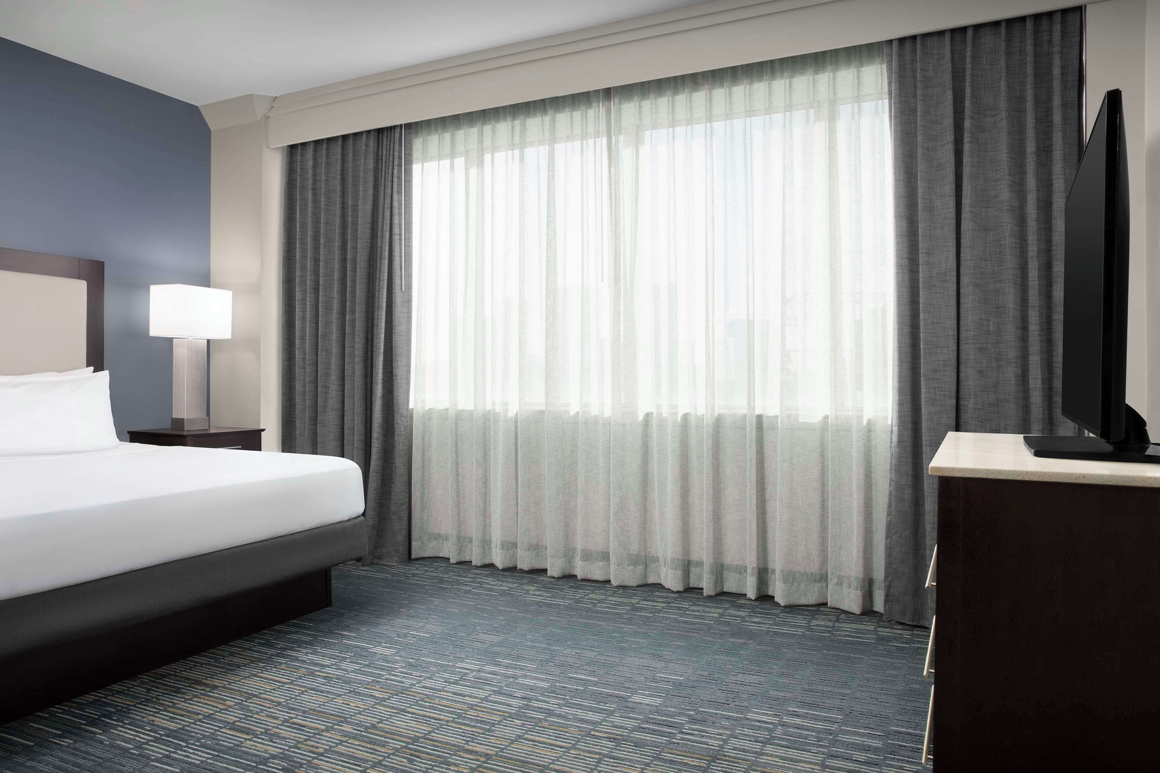 A hotel room with a bed, a desk, a television, and a large window with curtains.