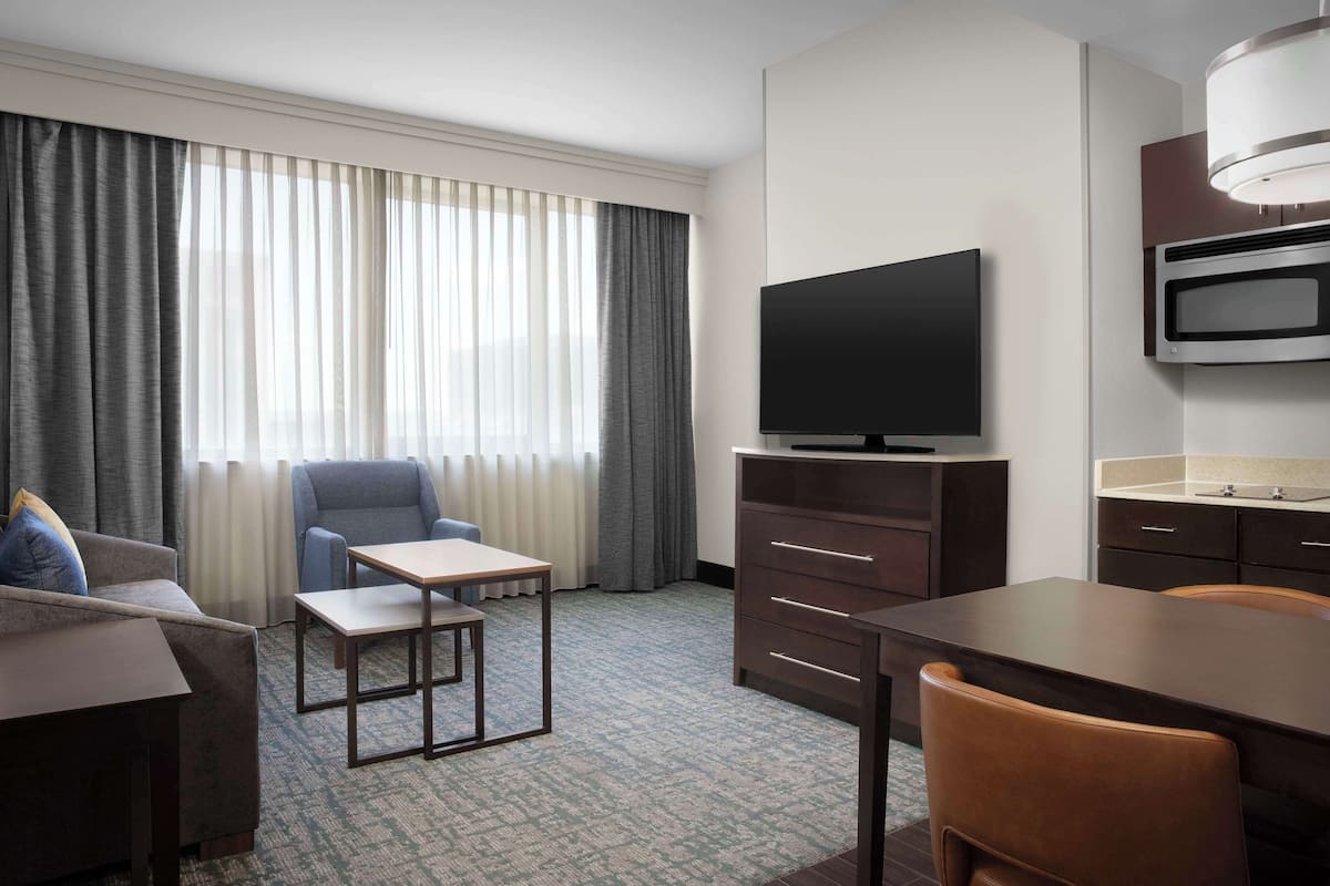 Habitación de hotel con sofá, sillón, mesa de comedor, televisor, minibar y un gran ventanal con cortinas.