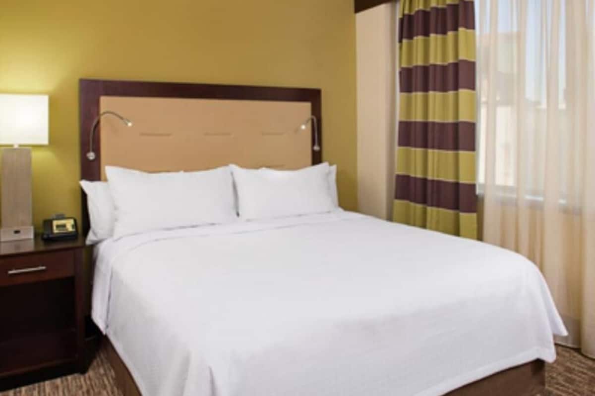 Una habitación de hotel con una cama grande, una mesita de noche con lámpara y una ventana con cortinas.