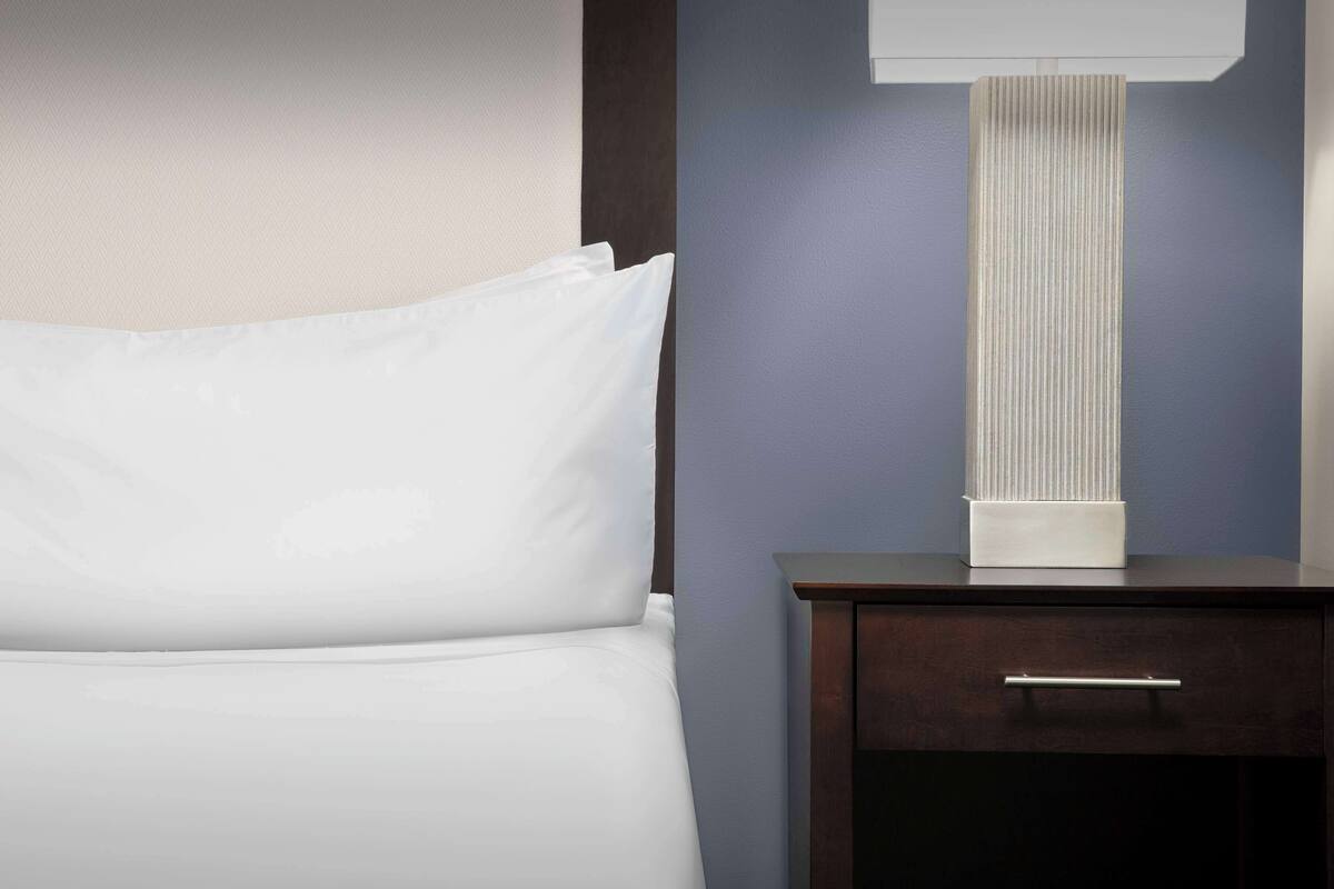 Una cama bien hecha con sábanas blancas, una mesita de noche de madera oscura con un cajón, y una lámpara de pie alta con textura en un tono azul.