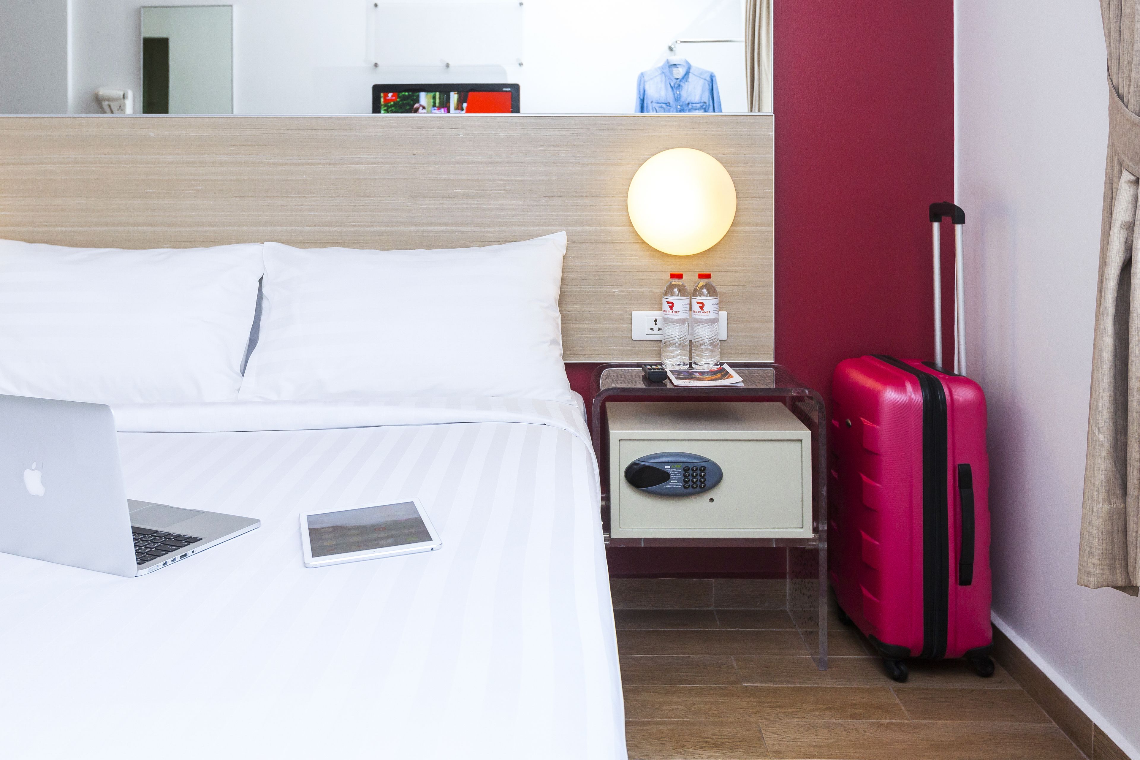 A hotel room with a bed, a nightstand with a lamp, a laptop, a smartphone, and a pink suitcase.