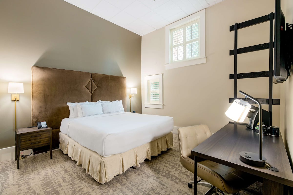 A hotel room with a large bed, bedside tables with lamps, a desk with a chair, and a window with blinds.