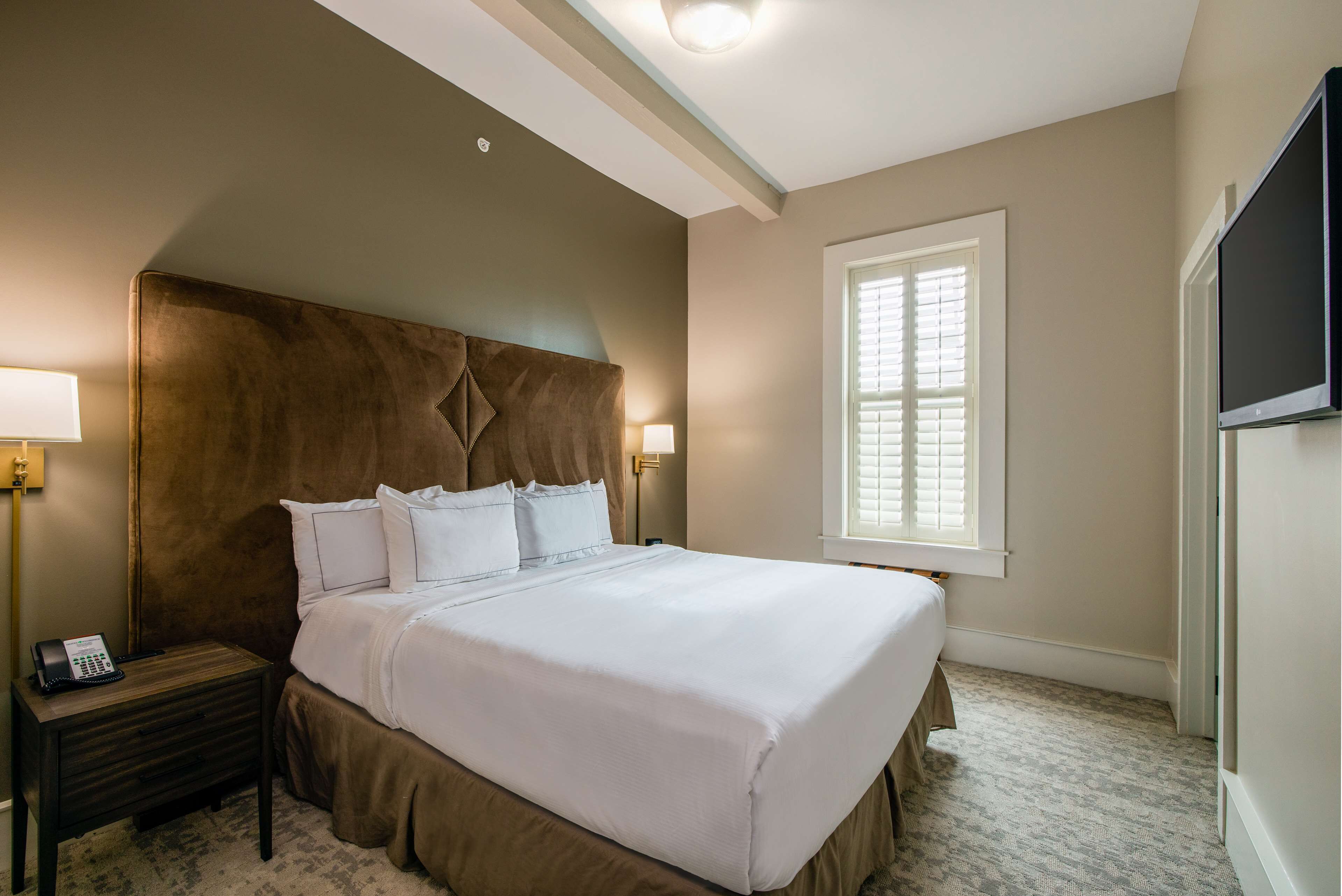 A neatly made bed with white linens, a wooden nightstand with a telephone, and a wall-mounted TV.