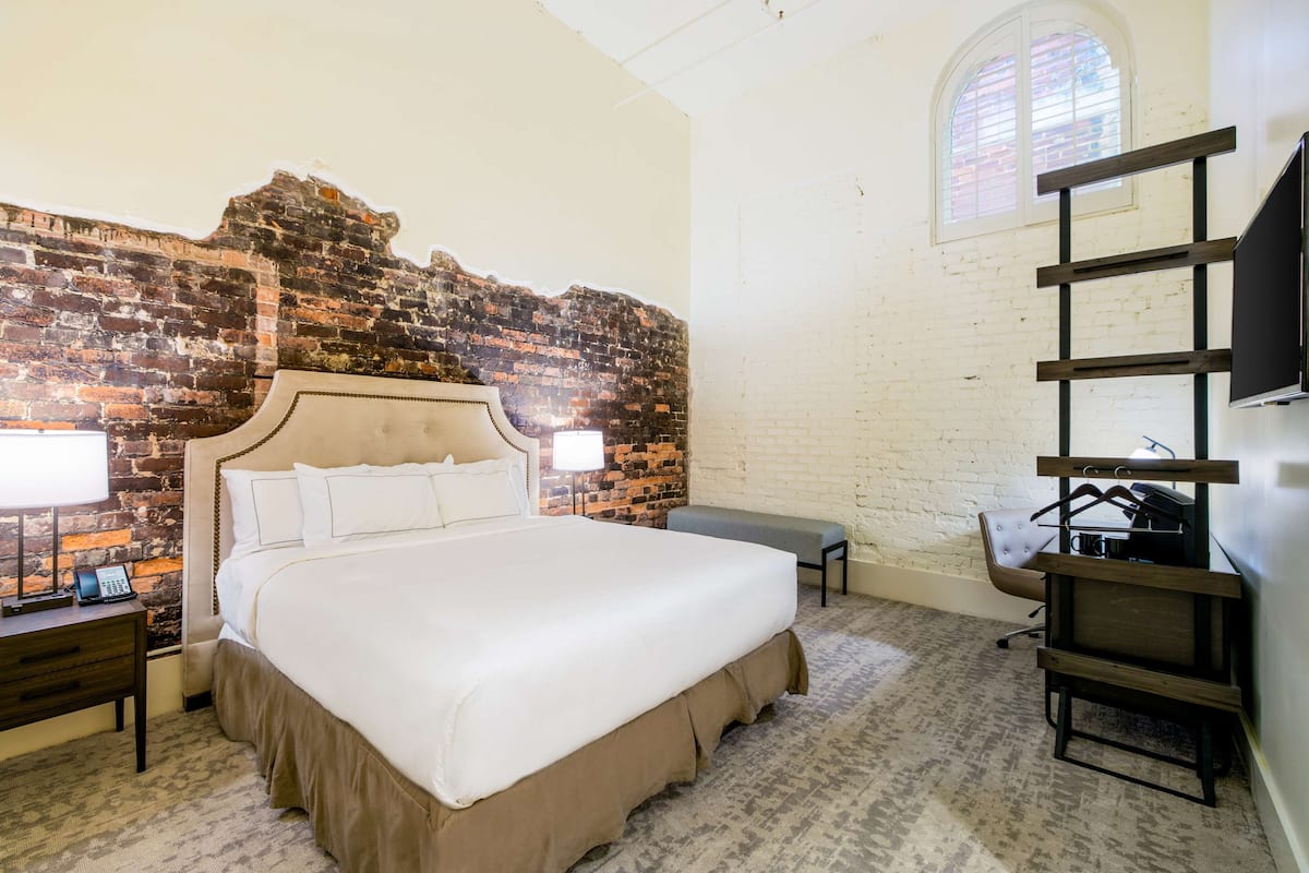 A hotel room with a brick wall, a large bed with white bedding, a bench, a desk with a lamp, and a TV mounted on the wall.