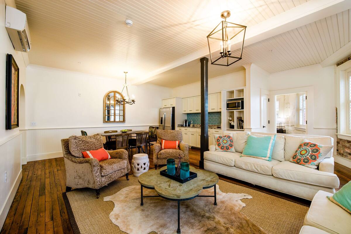 A spacious living room with a wooden floor, a white sofa with colorful cushions, two leopard print armchairs, a coffee table with decorative items, and a dining area with a wooden table and chairs.