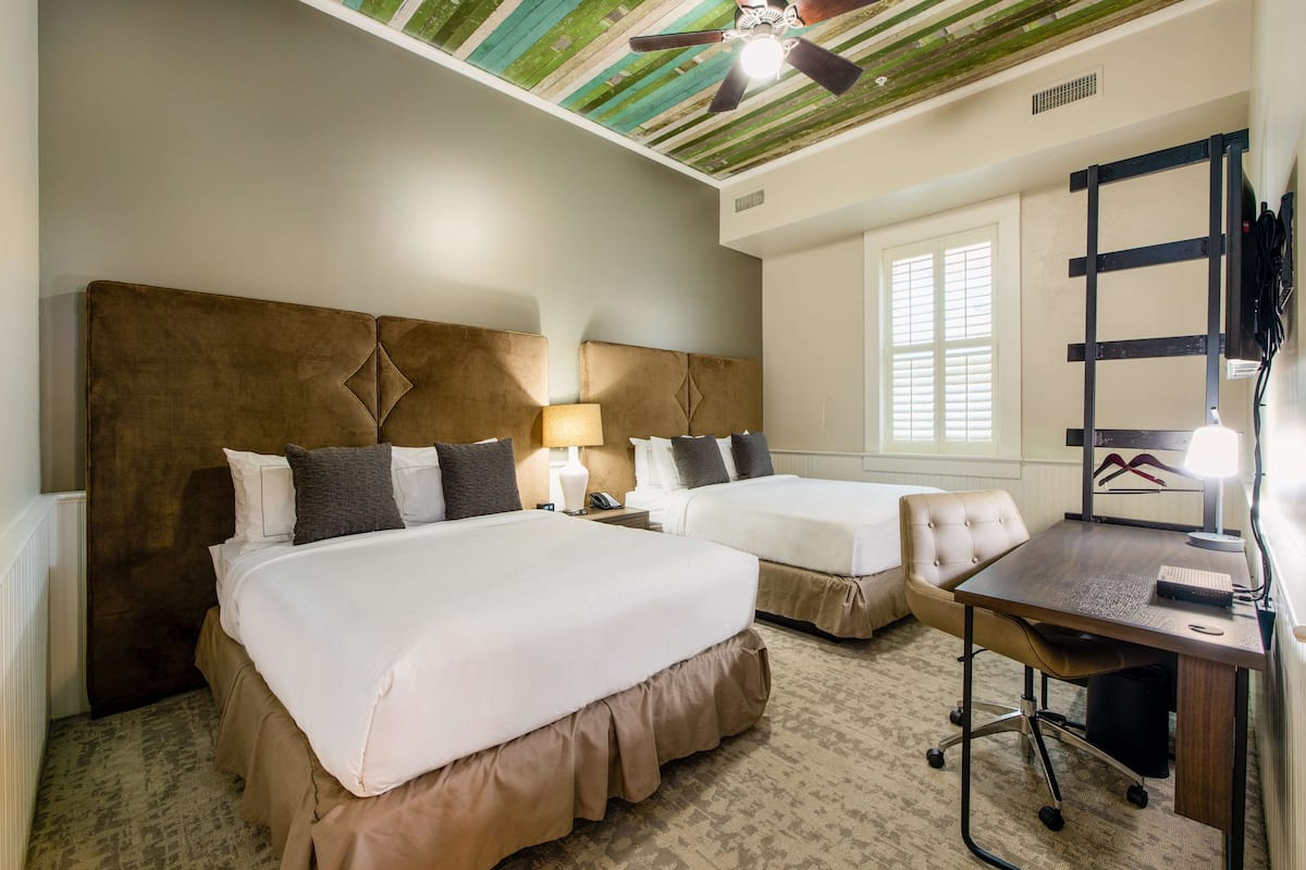 A hotel room with two beds, a desk, a chair, a ceiling fan, and a window with blinds.