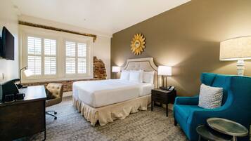A hotel room with a large bed, a desk, a blue armchair, and a small stool.