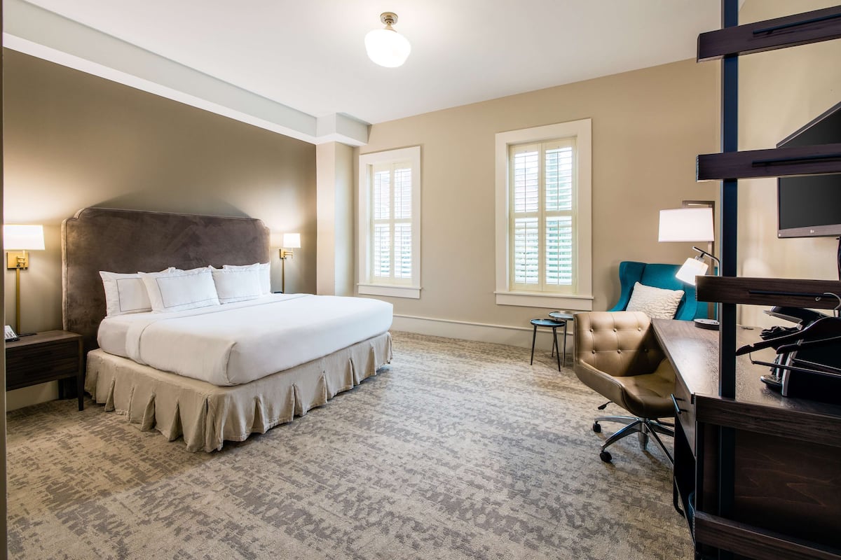 A hotel room with a large bed, a desk with a chair, a TV, and two windows with blinds.