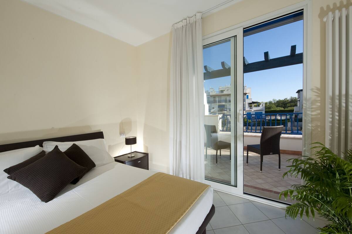A hotel room with a bed, a nightstand, a lamp, a balcony with a chair, and a view of buildings and greenery.