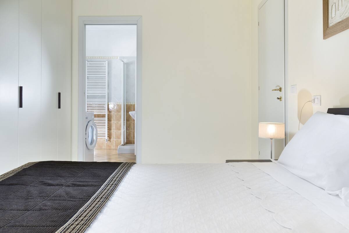 A hotel room with a bed, a nightstand with a lamp, a door leading to a bathroom, and a view of the bathroom.