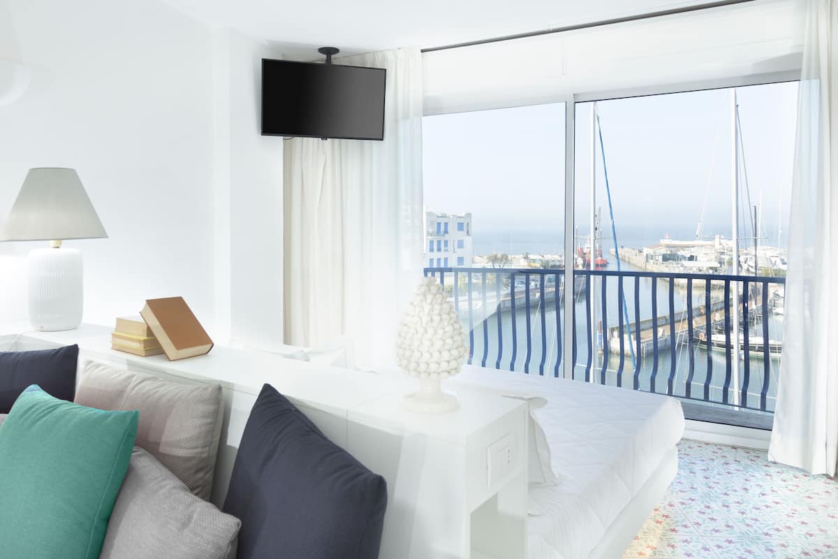 A hotel room with a bed, a TV, a table with books, a lamp, and a view of the sea and boats.