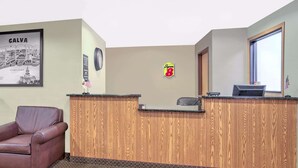 Un área de recepción con un mostrador de madera, un sillón de cuero, un cartel de un paisaje urbano enmarcado, un reloj y un letrero de Super 8.