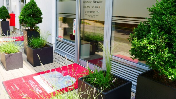 A hotel entrance with a red welcome mat, potted plants, and a sign indicating the name and office hours. - Parkhotel Marzahn (Berlin)