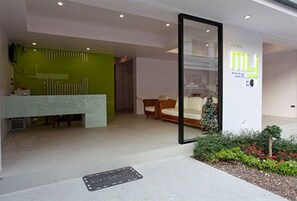 A modern interior with a reception desk, a green partition, a sofa, and a small plant area. - My Hotel (Patong)