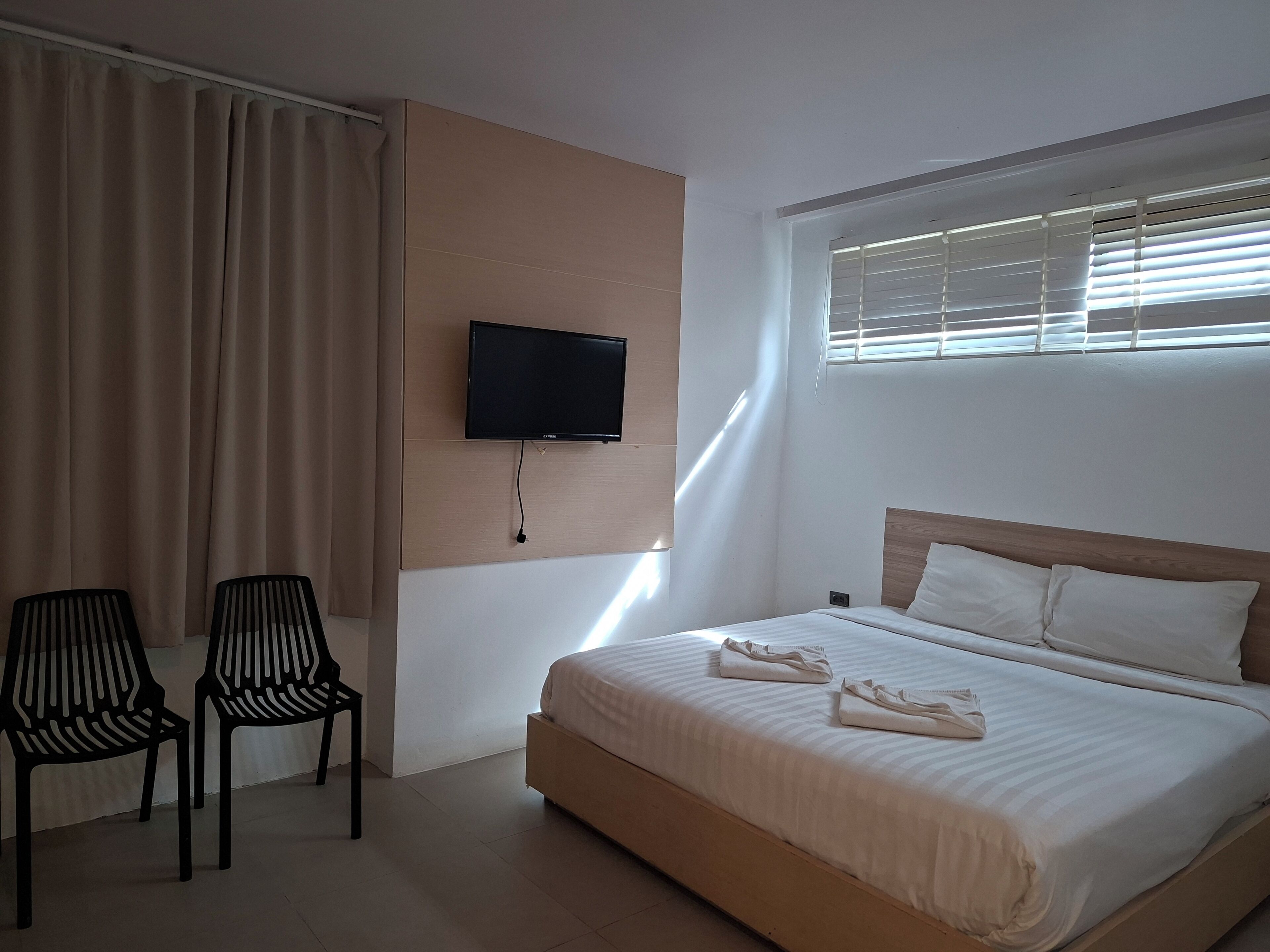 Quarto de hotel com cama, televisão na parede, duas cadeiras e janela com persianas.