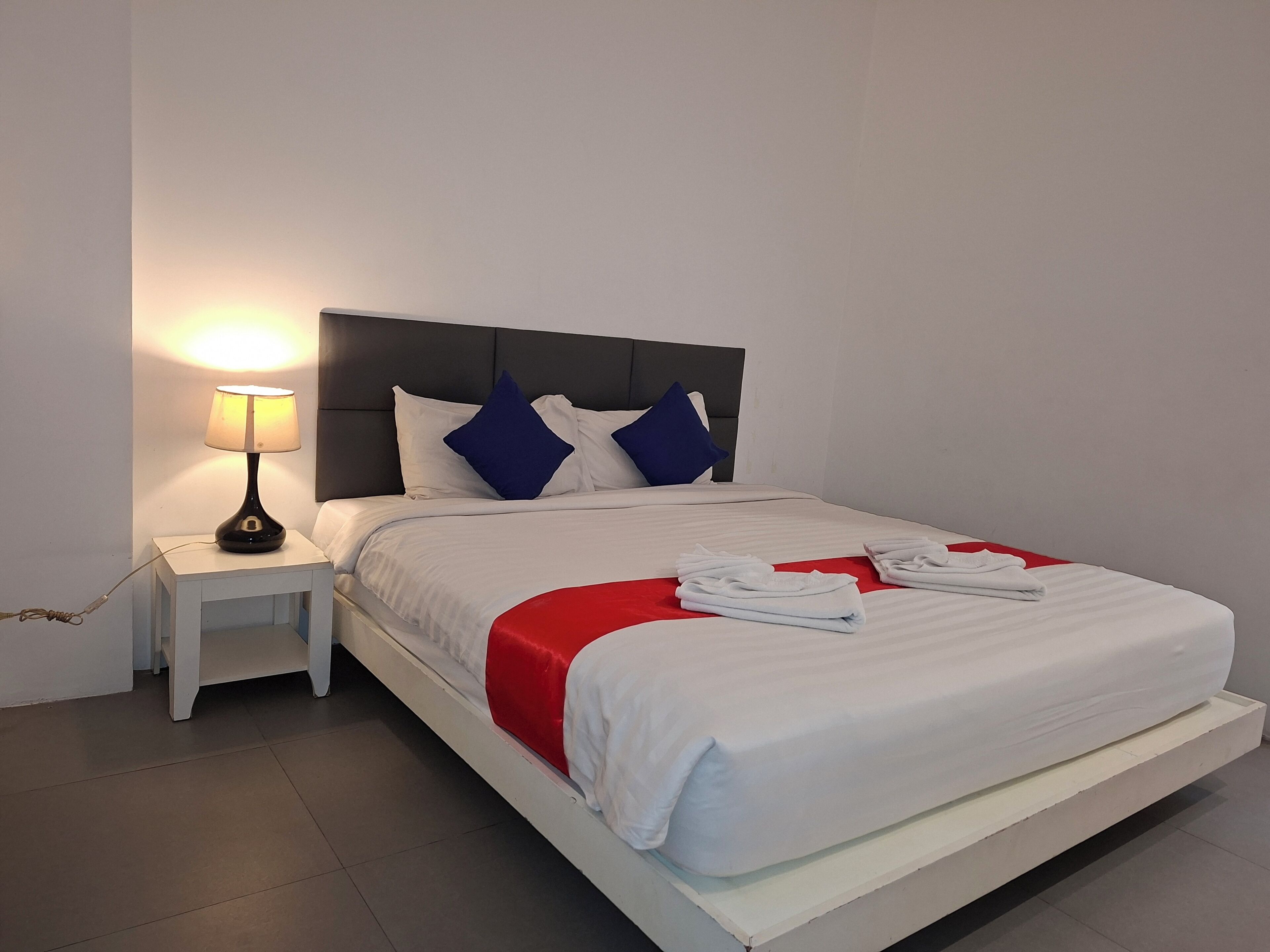 A neatly made bed with white bedding, a red accent, and blue pillows. There is a small bedside table with a lamp and a power cord.