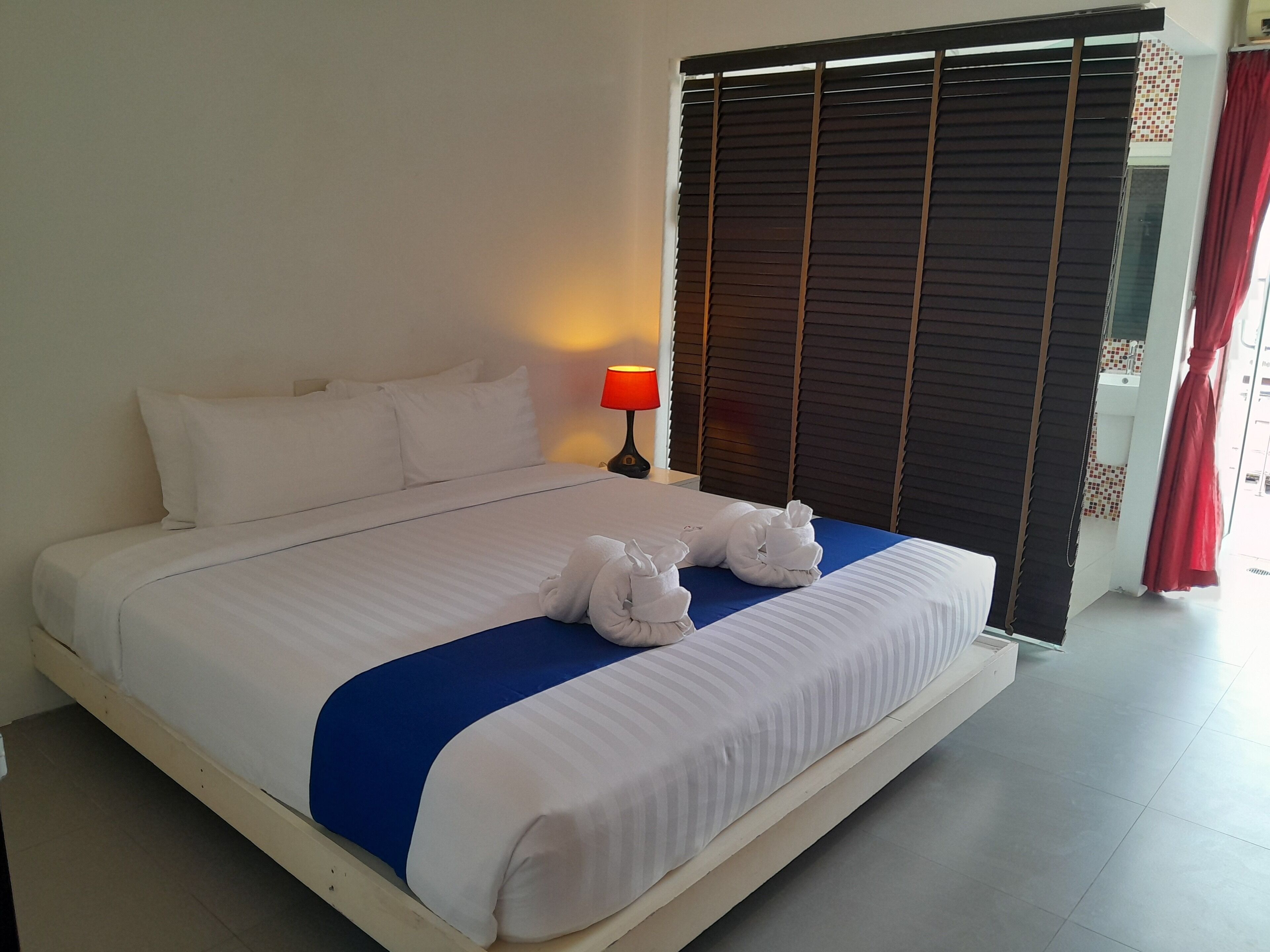 A neatly made bed with white and blue bedding, a red lamp on the nightstand, and a window with dark blinds.
