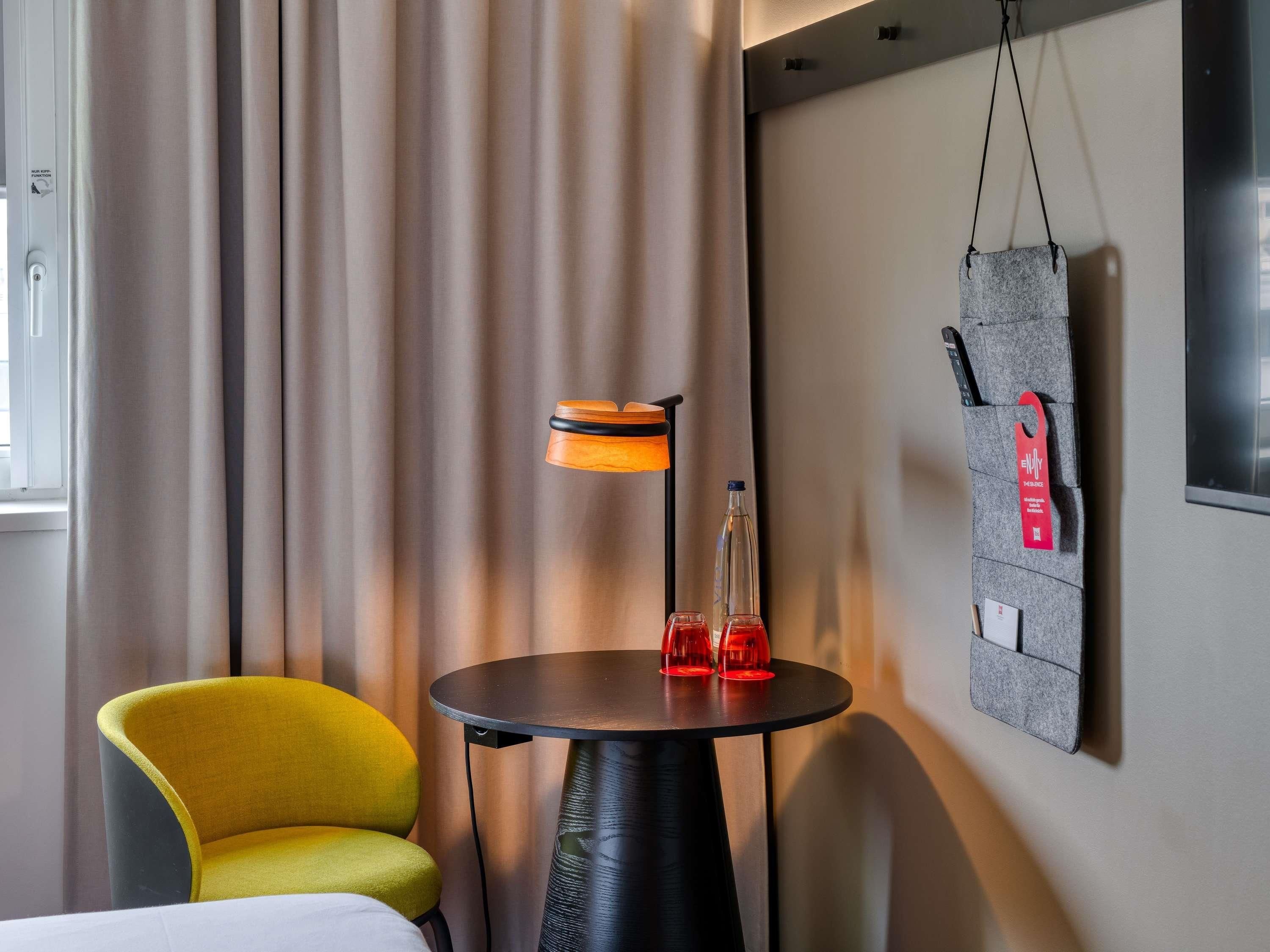 A hotel room with a yellow chair, a round table with two red glasses, a lamp, and a wall-mounted bag.