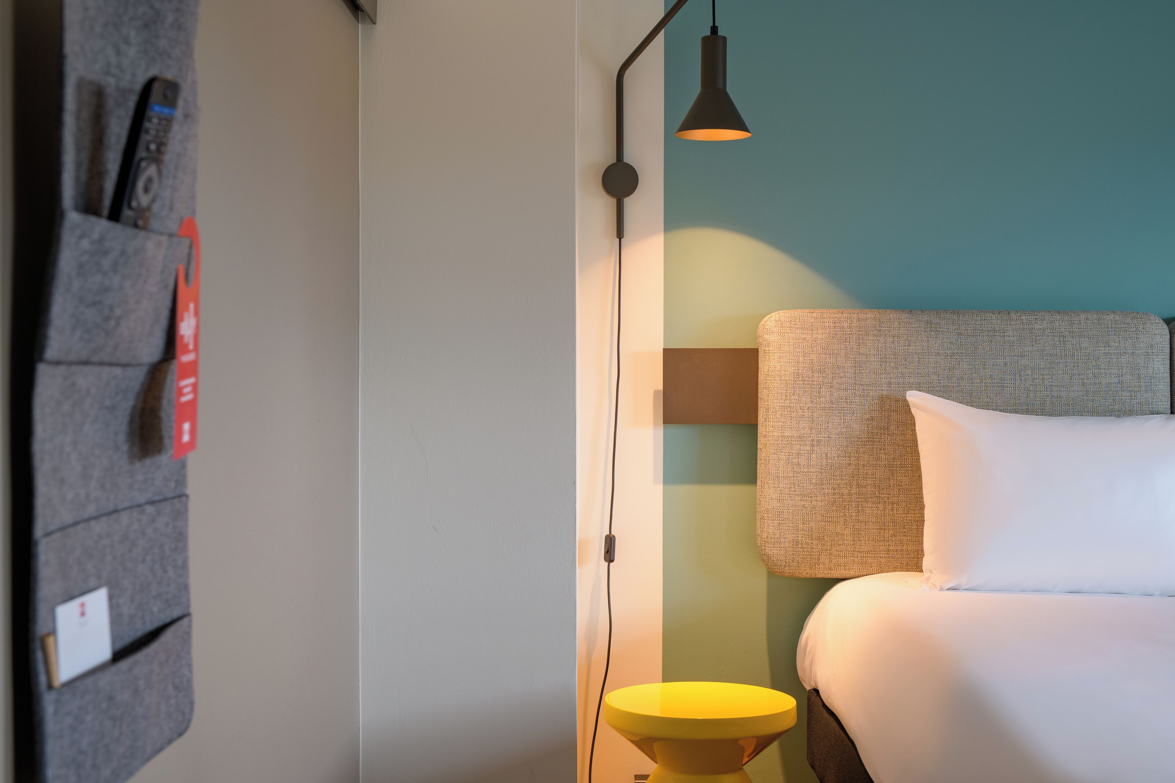 Une chambre d’hôtel bien rangée, avec un lit, une table de chevet, une lampe et une étagère fixée au mur, sur laquelle sont posés divers objets.