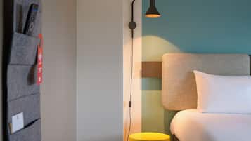 Une chambre d’hôtel bien rangée, avec un lit, une table de chevet, une lampe et une étagère fixée au mur, sur laquelle sont posés divers objets.