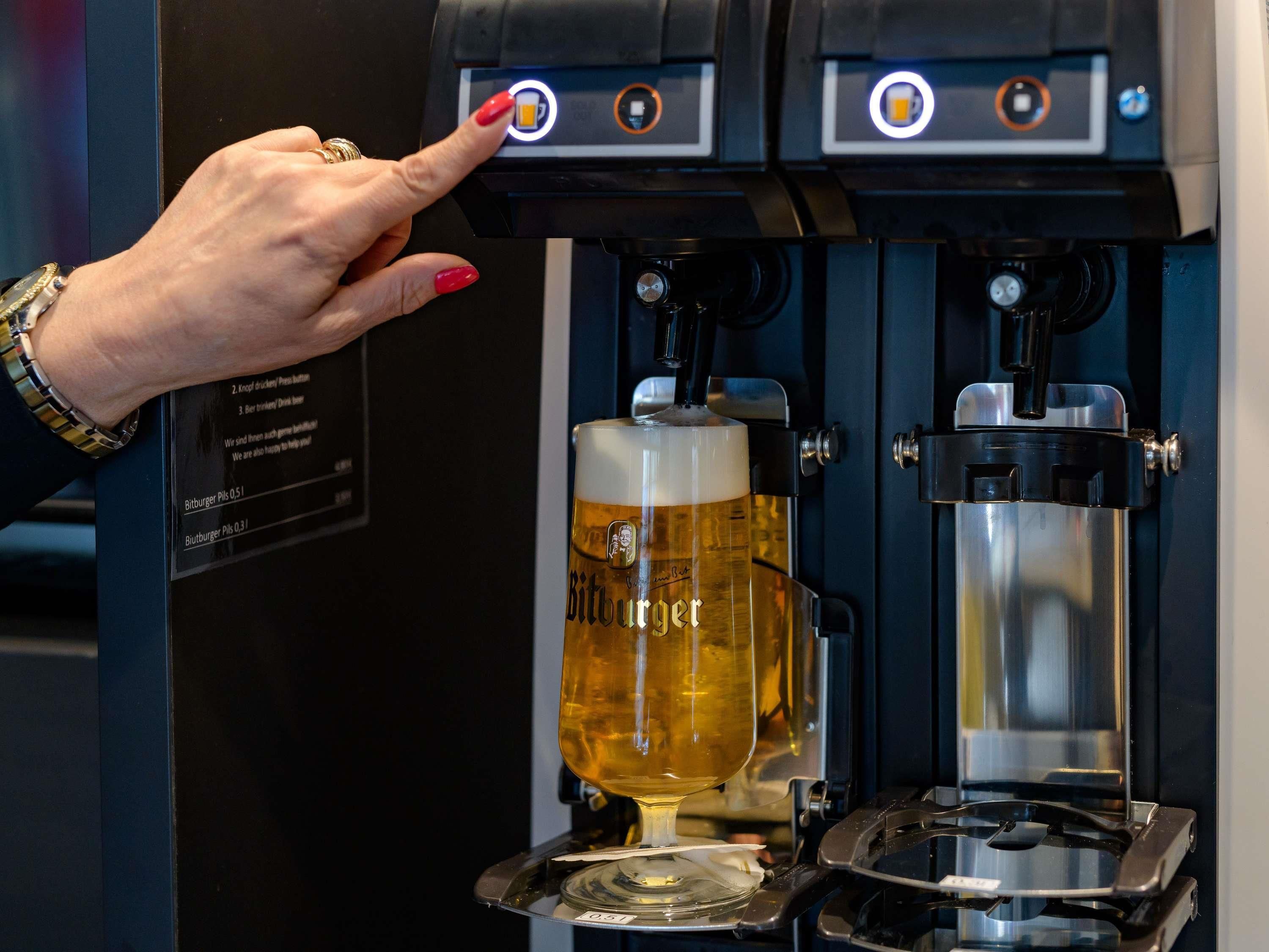 A hand pressing a button on a coffee machine, with a glass of beer and a coffee pot visible.