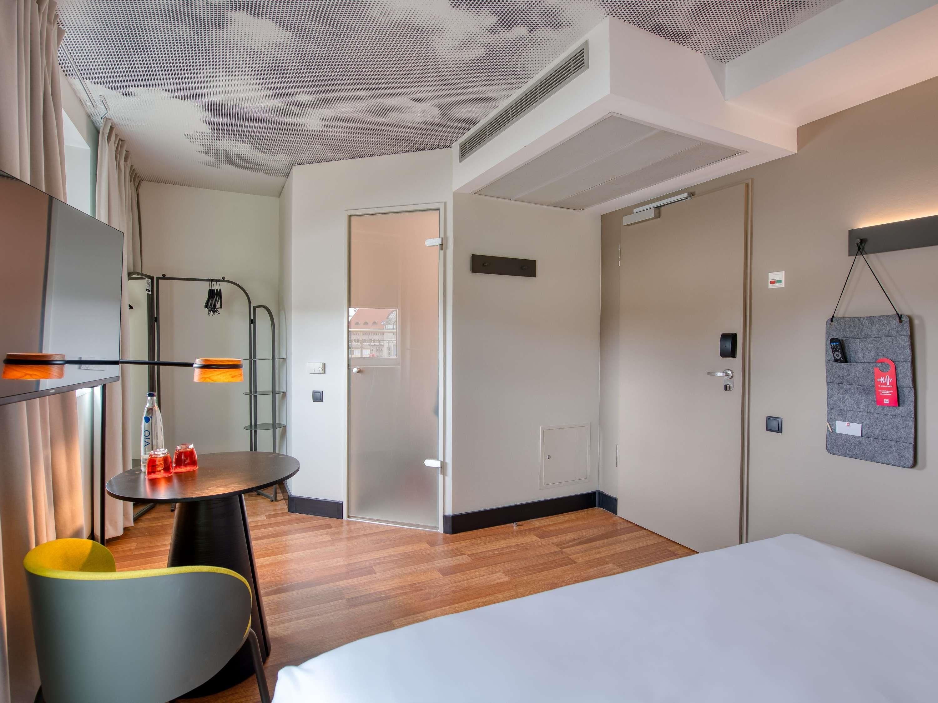 Una habitación de hotel moderna con una cama, una pequeña mesa redonda con sillas, un armario y una estación de carga en la pared.