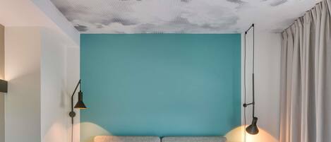 Habitación de hotel moderna con una cama grande, dos lámparas de noche, una mesita redonda y vistas del cielo a través de una ventana con cortinas.