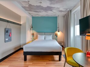 A modern hotel room with a bed, bedside tables, a TV, a chair, and a unique ceiling design. - ibis Berlin Kurfürstendamm (Berlin)