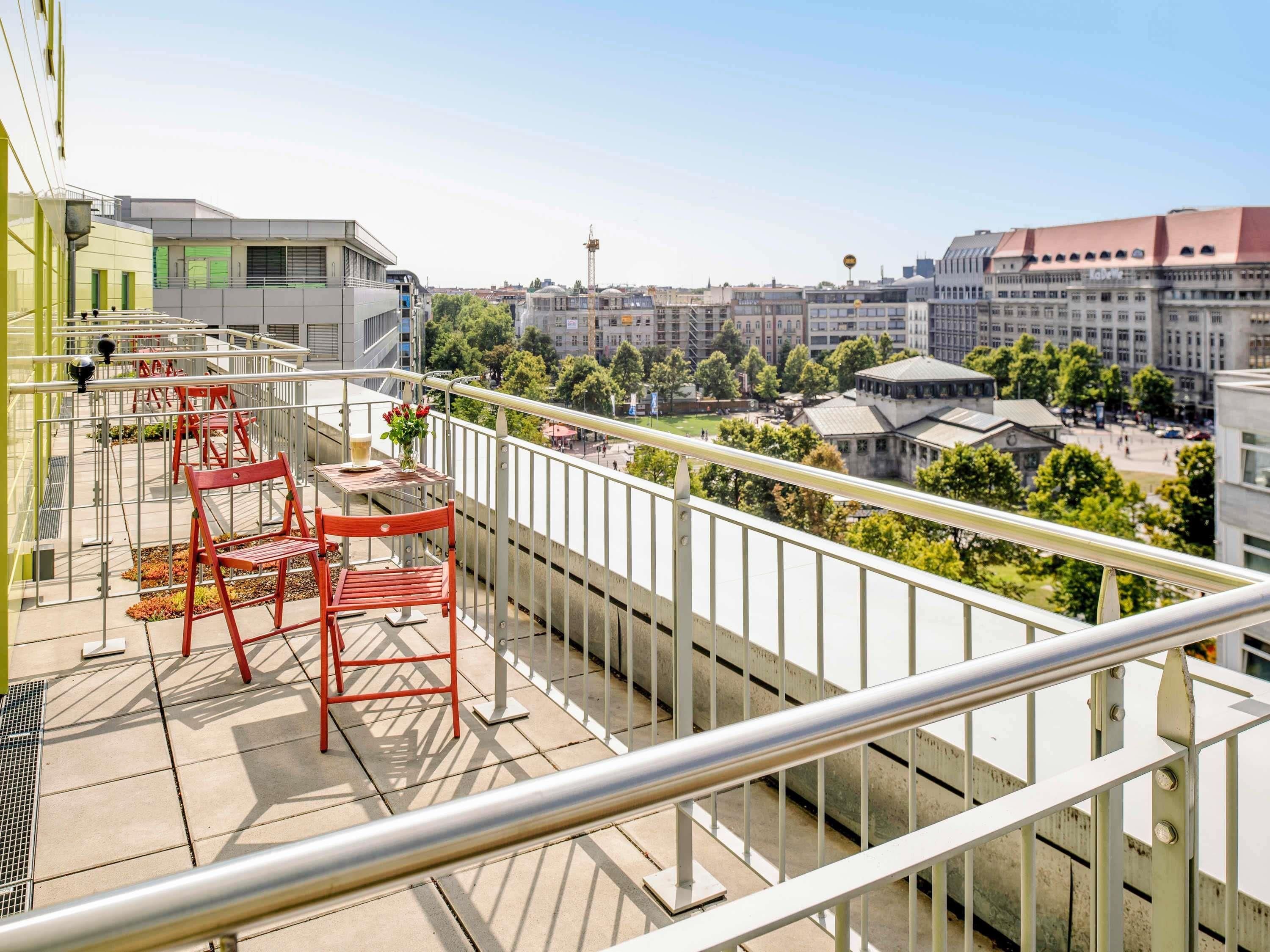 A balcony with red chairs and a table, overlooking a cityscape with buildings and a tower.
