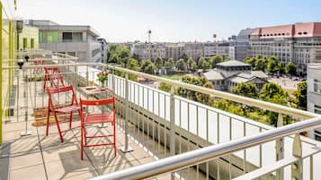 A balcony with red chairs and a table, overlooking a cityscape with buildings and a tower.