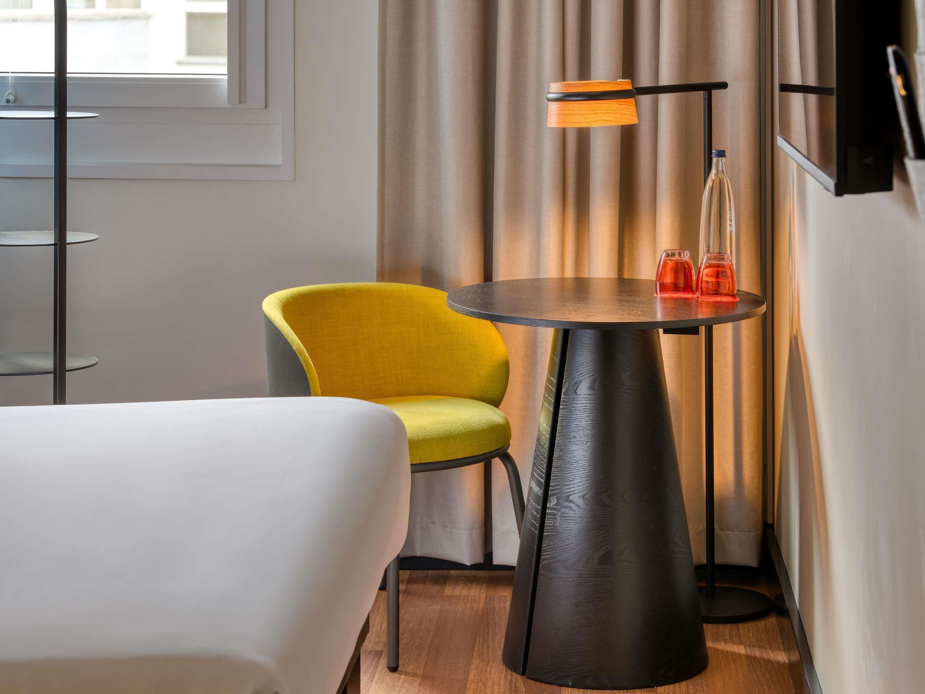 A modern hotel room with a round table, a yellow armchair, a white sofa, and a floor lamp.