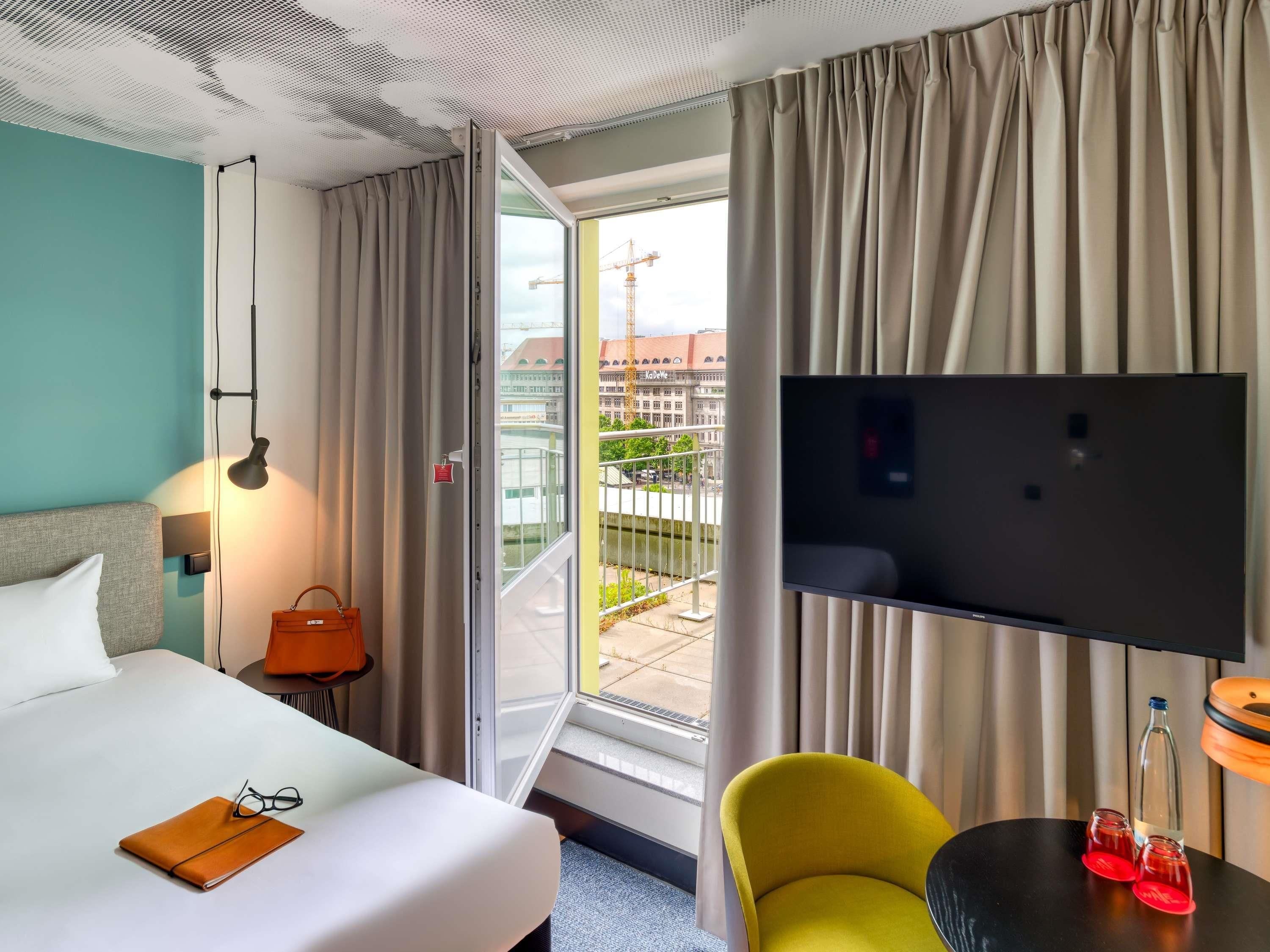 Ein Hotelzimmer mit einem Bett, einem gelben Sessel, einem Fernseher und einem Balkon mit Blick auf Gebäude.