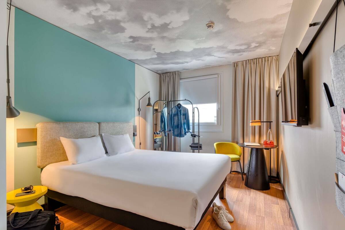 A hotel room with a bed, a desk, a chair, a coat rack, and a TV.