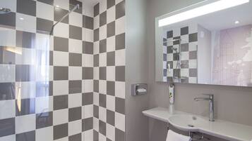 A modern bathroom with a checkered tile wall, a glass shower enclosure, a white sink with a mirror, and a dark wood floor.