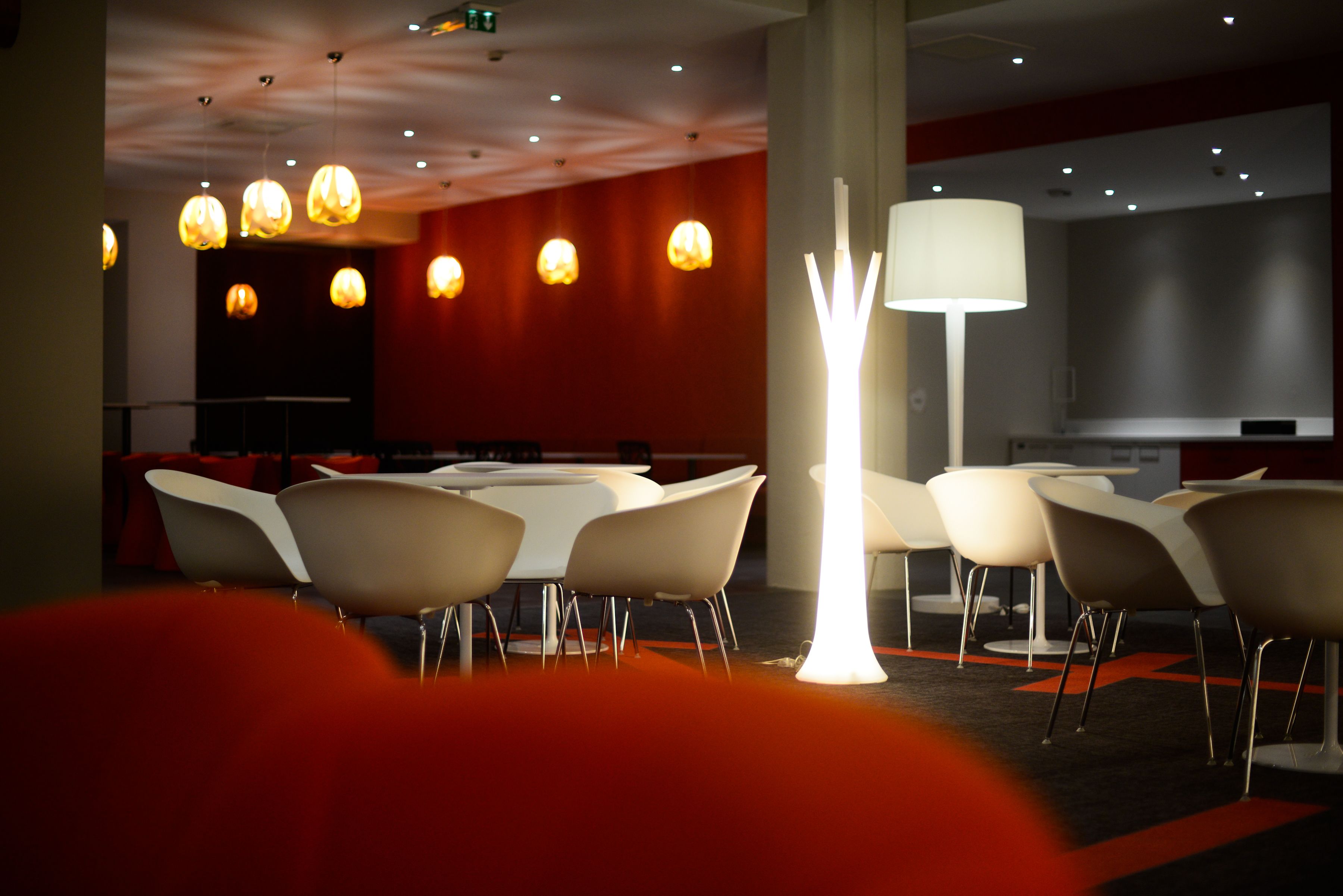 Un comedor moderno con mesas y sillas blancas, una lámpara de pie alta y luces colgantes.