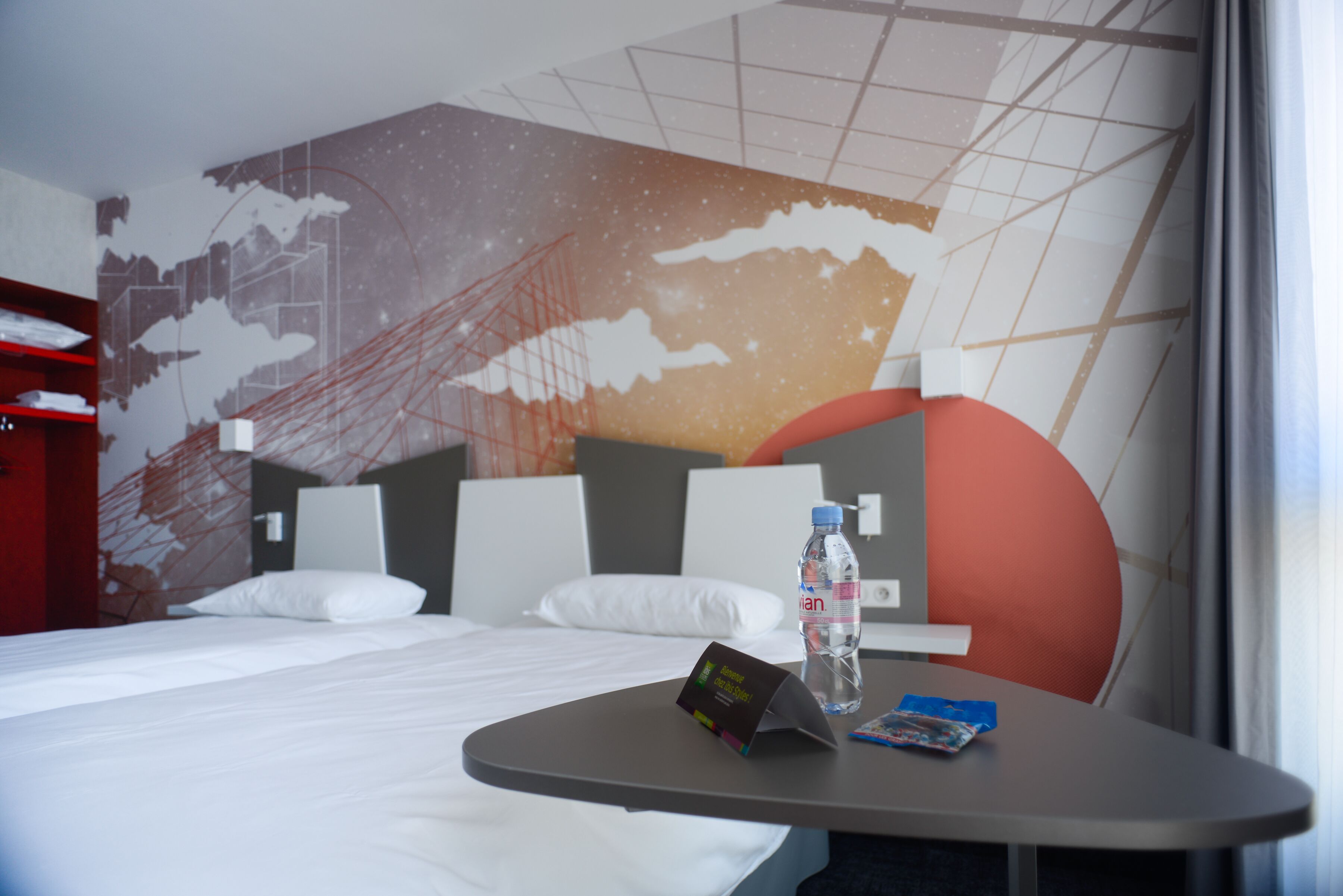 Ett hotellrum med en säng, ett bord med en flaska och ett kort, och en vägg prydd med en väggmålning som föreställer en stadsbild.