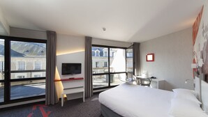 Habitación de hotel con una cama grande, un escritorio, una silla, un televisor y un balcón con vistas a edificios.