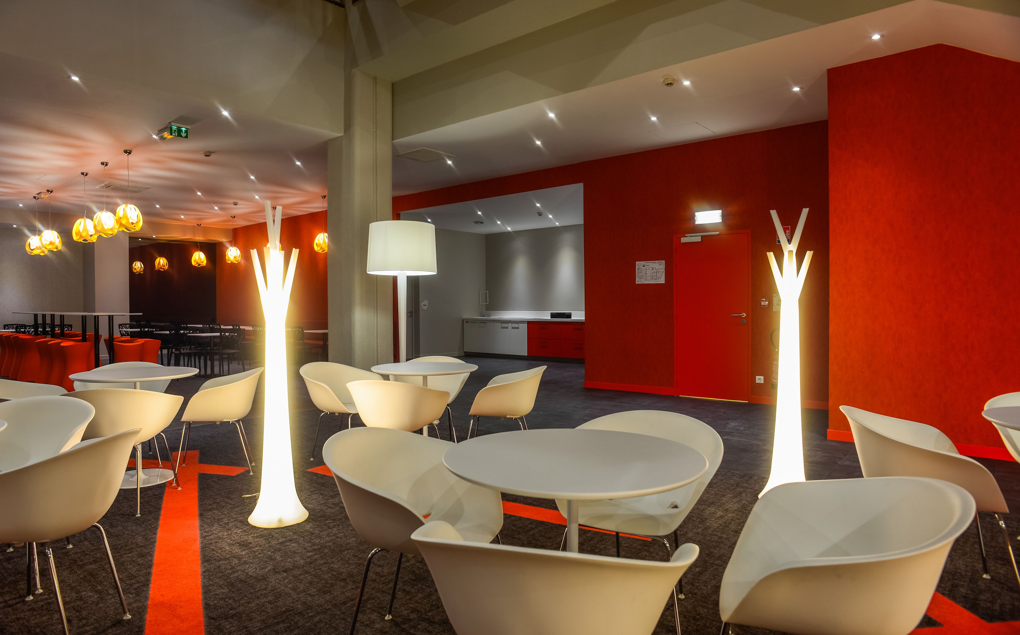 Un lobby de hotel moderno con decoración blanco y rojo, varias zonas de estar y una cocina visible al fondo.